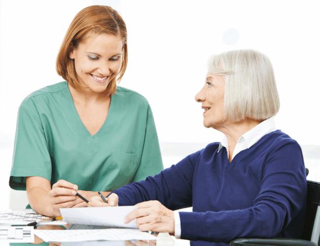 senior woman in a wheelchair smiling up at a nurse while filling out a crossword puzzle