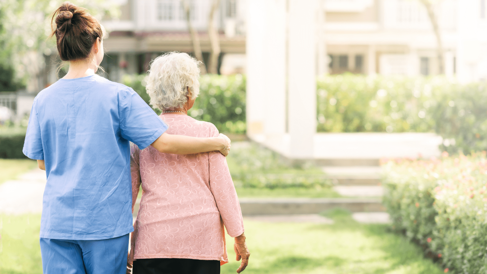 Caregiver with arm around the shoulders of a senior resident walking toward a building.