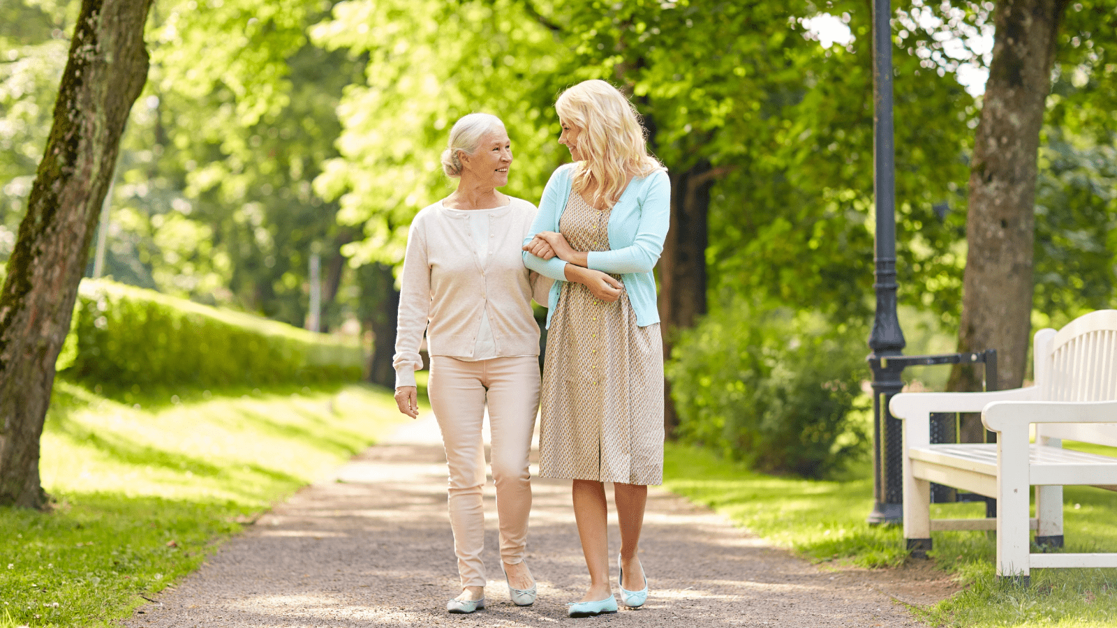 A mother and daughter walk down a park path with trees and a bench.