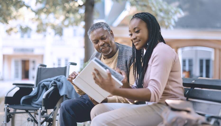 younger woman going through a book with a senior man outside on a park bench