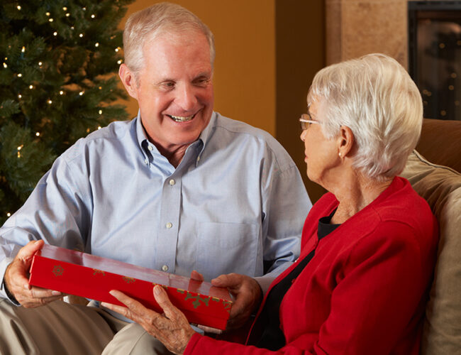 A man and a woman sit on a couch exchanging a holiday gift