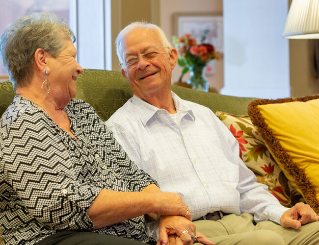 A couple seated on a sofa looking at each other smiling.