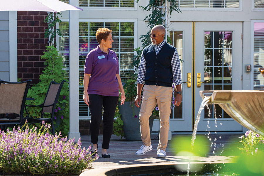 A caregiver and resident walk outdoors with a fountain in the foreground.