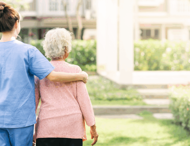 Caregiver with arm around the shoulders of a senior resident walking toward a building.