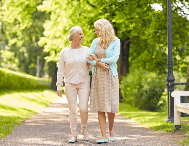 A mother and daughter walk down a park path with trees and a bench.