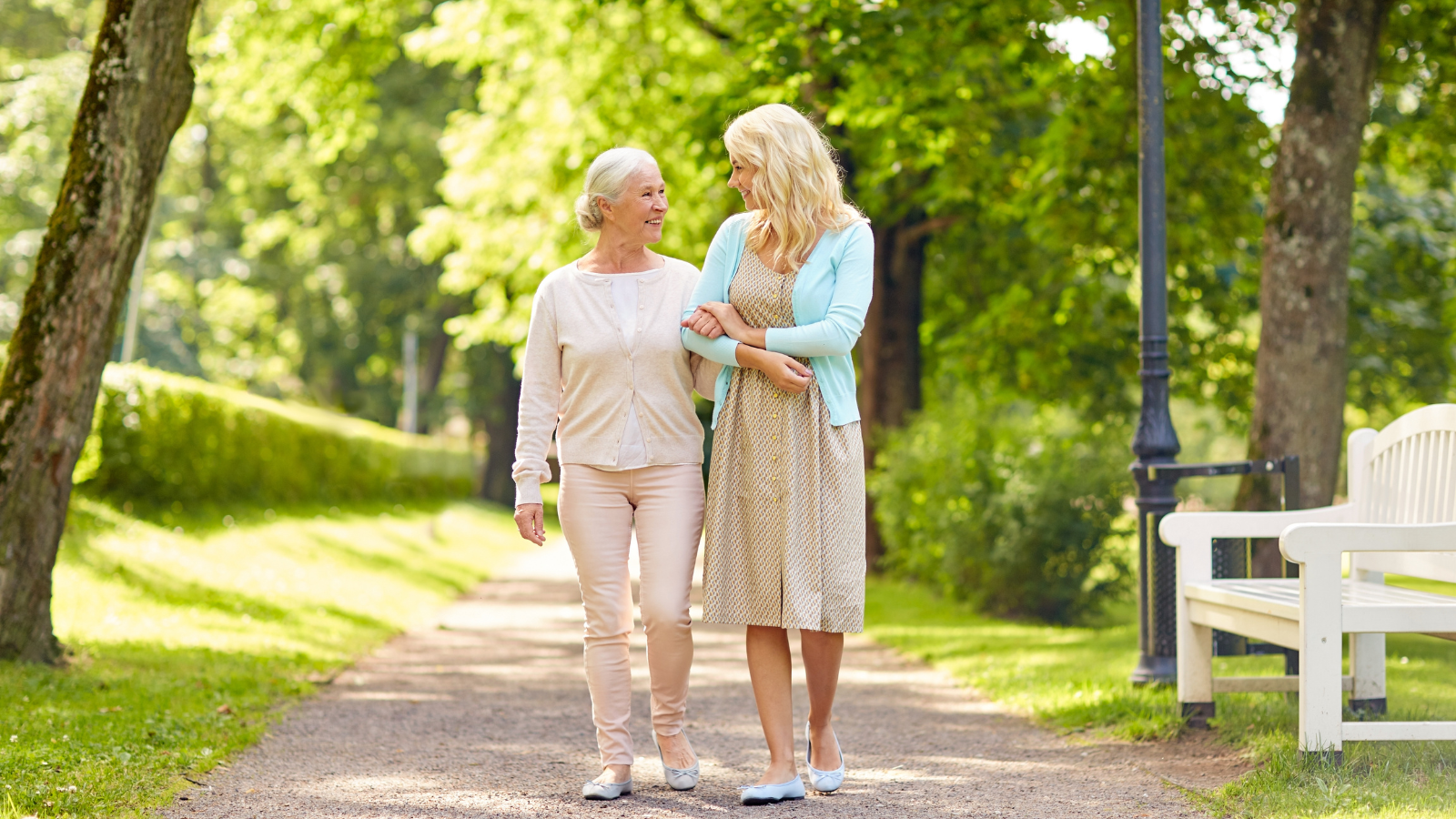 A mother and daughter walk down a park path with trees and a bench.